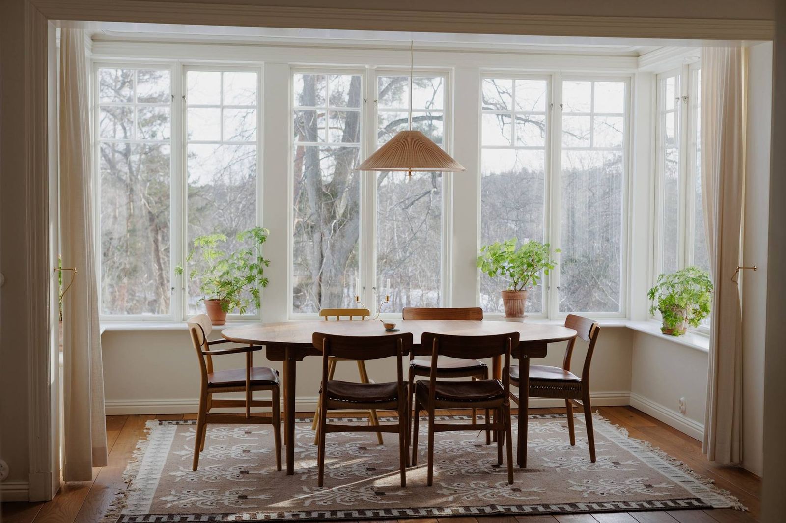 Dining room with oval wood table, mid-century chairs and wraparound windows onto bare winter trees